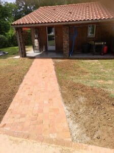 Allée de pavés en terre cuite menant à une maison avec toit de tuiles rouges et piliers en pierre naturelle.