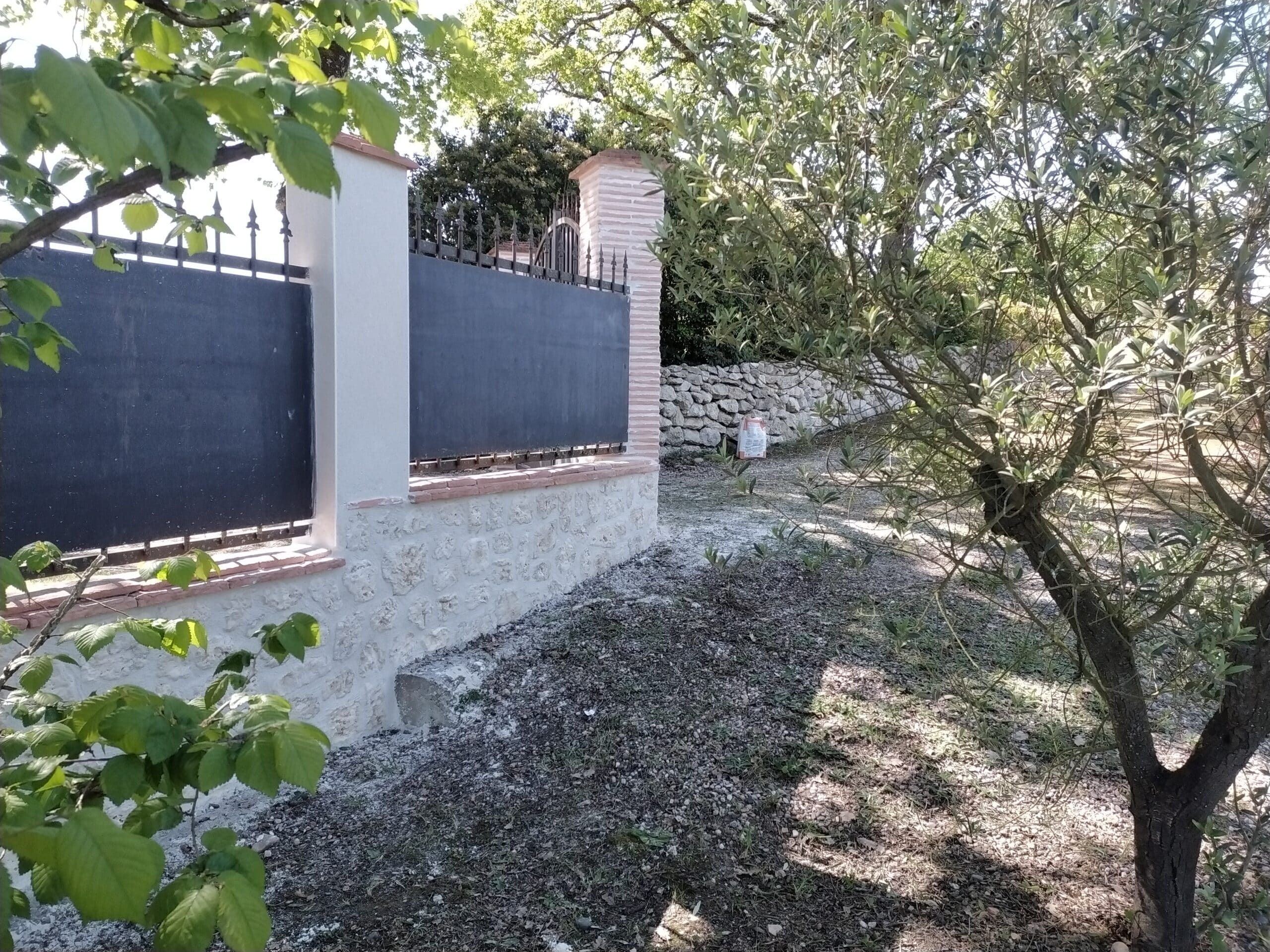 Clôture élégante en pierre, crépi blanc et fer forgé noir bordée par un olivier et des feuillages dans un jardin ensoleillé.