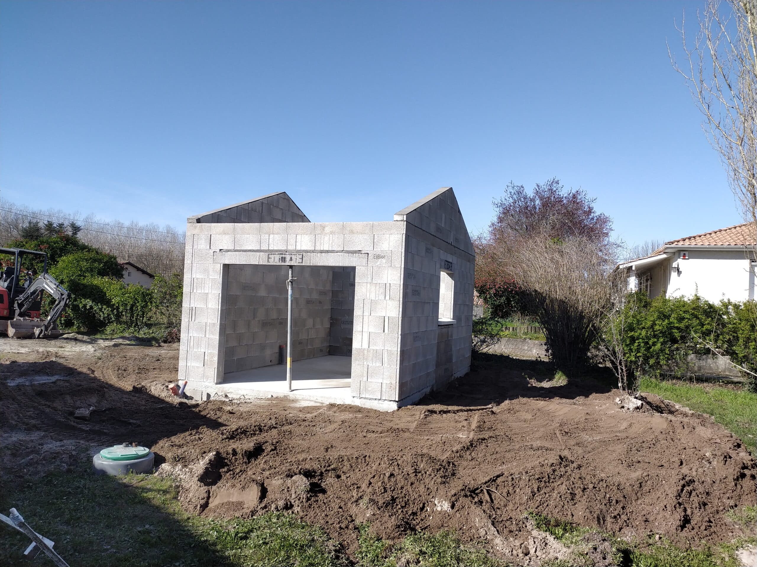 Chantier de garage en parpaings avec pelleteuse sur terrain en terre sous un grand ciel bleu.