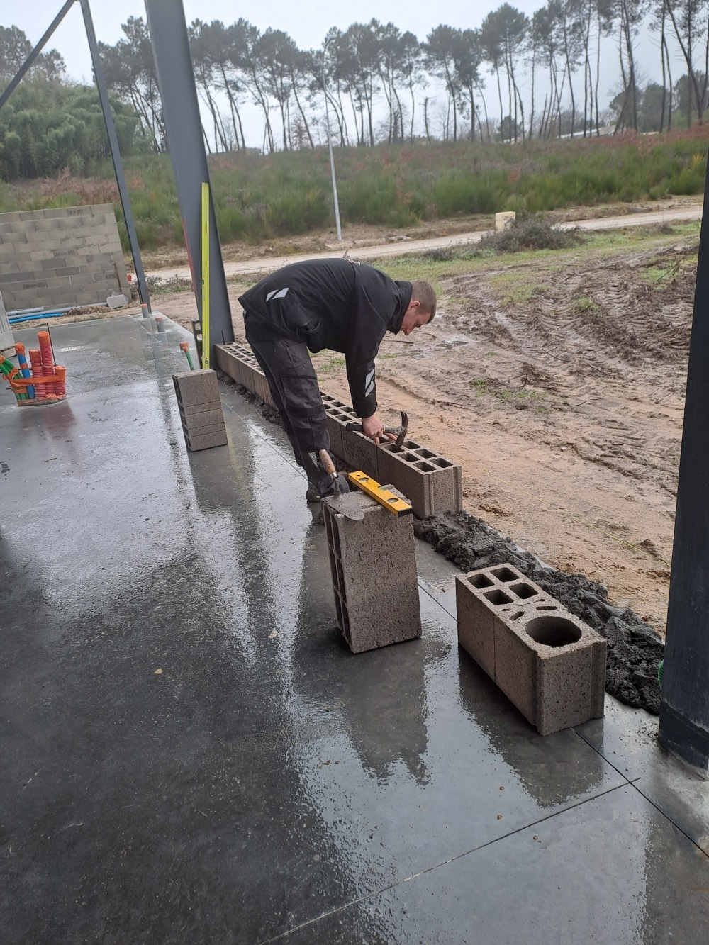 Maçon posant des parpaings sur une dalle humide avec un marteau et un niveau lors d'un chantier de construction.