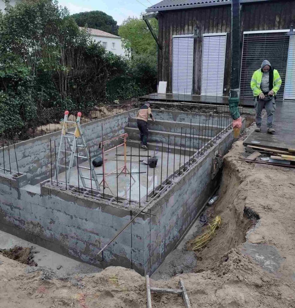 Ouvriers sur le chantier de construction d'une piscine en blocs de béton avec fers à béton apparents dans un jardin.