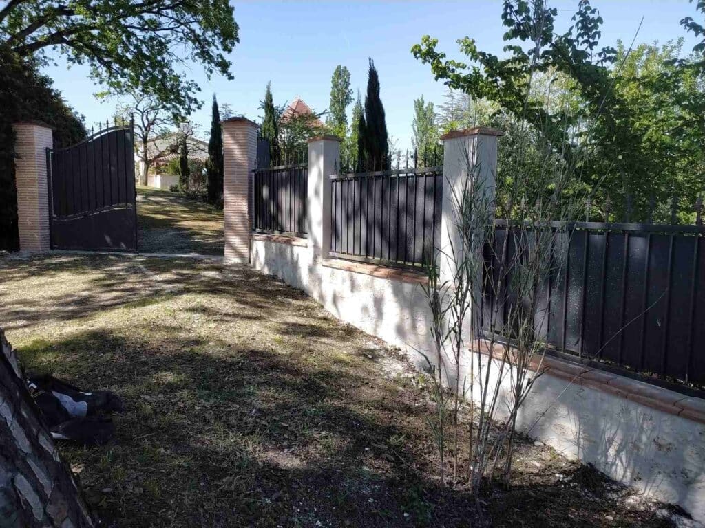 Portail noir ouvert sur un jardin avec clôture en fer forgé et muret blanc sous un ciel bleu.